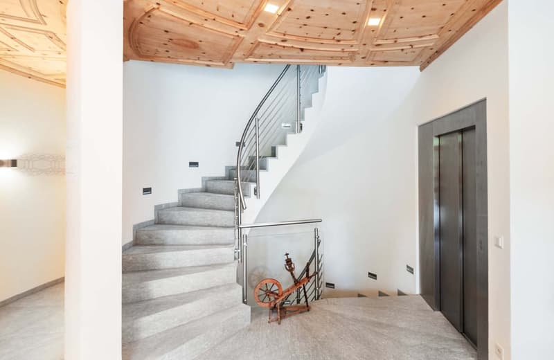 The image shows the interior of a building with a spiral staircase made of concrete and metal railings. The ceiling has a wooden design with recessed lighting. There is a fireplace visible in the corner of the room. The walls are white and the floor is ma