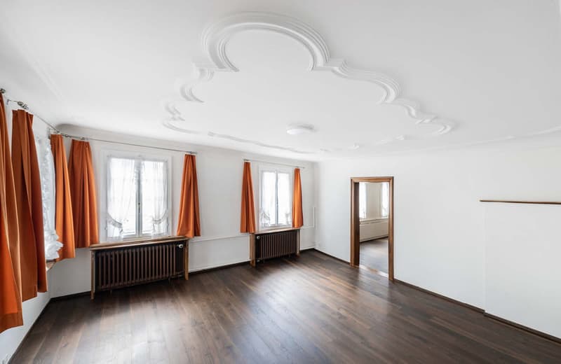 Spacious living room with hardwood floors, white walls, large windows, ornate ceiling molding, and a fireplace with radiators below the windows.