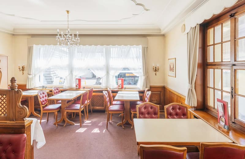 This image shows a traditional, cozy dining room with wooden furniture and decor. The room has large windows that let in natural light, a crystal chandelier, and red tufted leather chairs around wooden tables. The overall atmosphere appears to be that of