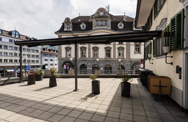 The image shows a historic, ornate building with a distinctive roofline and arched windows. The building has a grand, classical facade with decorative architectural elements. In front of the building, there is a paved plaza with potted plants and some out