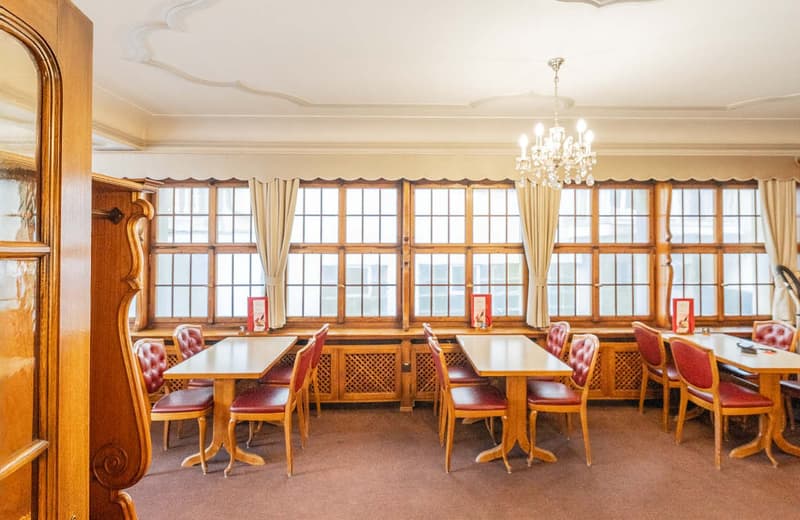 Large dining room with wooden furniture, including a long table with red upholstered chairs, a chandelier, and large windows providing natural light.