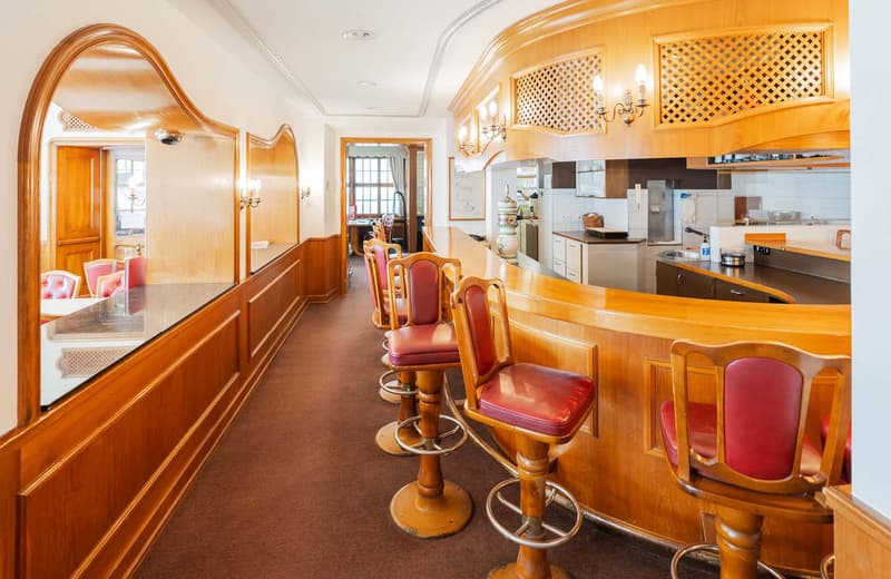 The image shows a cozy and vintage-style kitchen and dining area. The space features warm, wooden tones with intricate lattice-patterned accents on the walls and cabinets. There is a curved bar-style counter with red leather stools, as well as a dining ta