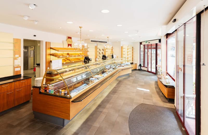 Large open bakery/cafe space with wooden shelves, display cases, and counters displaying various baked goods and pastries. The space has high ceilings, recessed lighting, and a glass storefront entrance.
