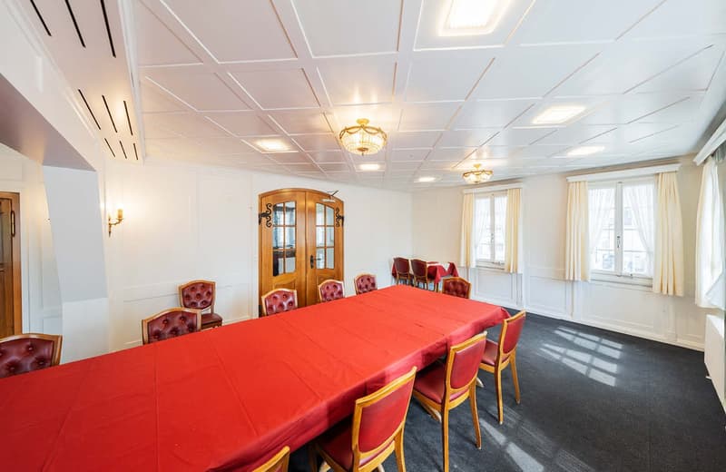 Large meeting room with a long red table, wooden chairs with red upholstery, wooden doors with glass panels, and a tiled ceiling with recessed lighting.