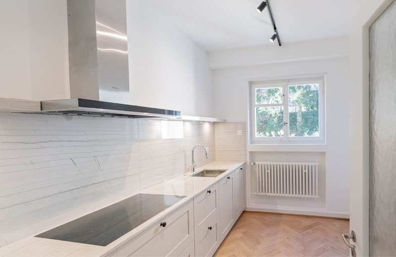 modern kitchen with white cabinetry, marble countertop, undermount sink, stainless steel range hood, induction cooktop, radiator heater, wooden floor