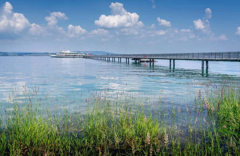 Scenic view of a long wooden pier extending over a calm lake or bay, with a passenger boat docked at the end. The water is a beautiful turquoise color, and the surrounding landscape includes lush green vegetation along the shoreline.