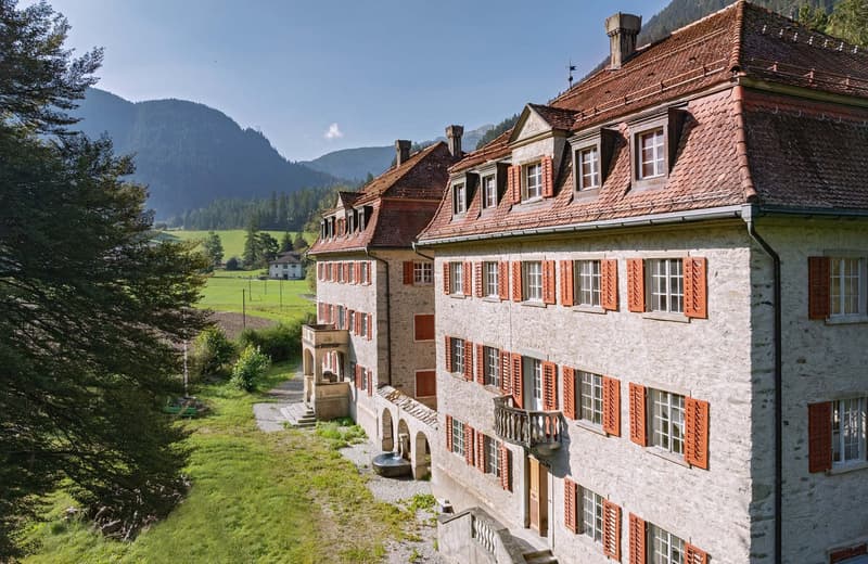 A multi-story stone building with red tiled roofs, surrounded by a lush green landscape and mountains in the background. The building has several balconies and windows with red shutters.