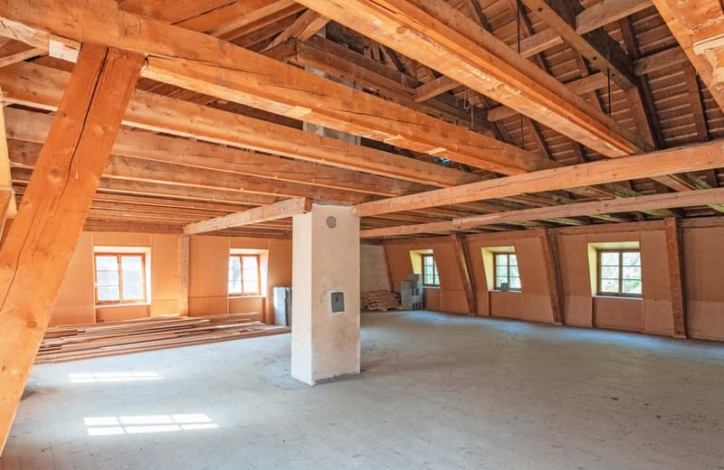 The image shows the interior of a large, open space with a wooden ceiling and exposed wooden beams. The room has several large windows that let in natural light. The floor appears to be concrete or a similar material. The space appears to be in a state of