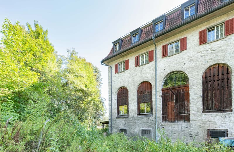 A historic stone building with red-tiled roofs, wooden shutters, and arched doorways surrounded by lush greenery and trees