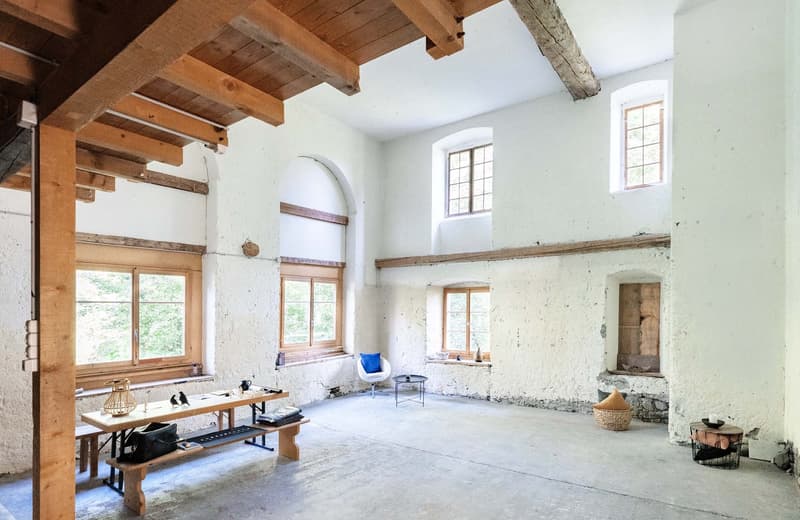 Large open-plan living space with exposed wooden beams, white walls, arched windows, and a fireplace. The room has a rustic, industrial feel with a wooden table, chairs, and other furnishings.