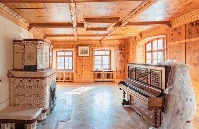 Spacious room with wooden walls and ceiling, large windows, ornate fireplace, and a grand piano covered in a white sheet