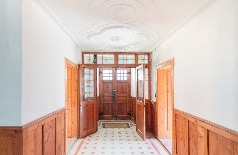 The image shows a spacious entrance hall with a high ceiling and ornate plaster moldings. The walls are lined with wood paneling, and the floor is covered in a patterned tile. The main entrance features a set of large, wooden double doors with stained gla