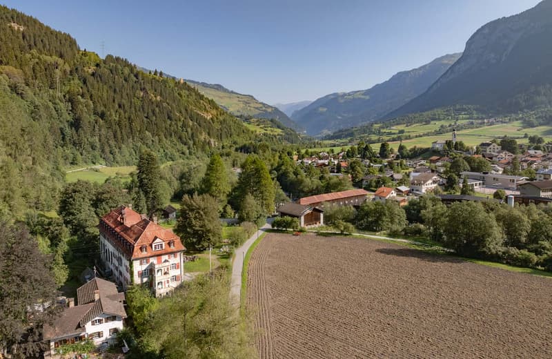 The image shows a picturesque mountain landscape with a small town nestled in the valley. The town is surrounded by lush green forests and fields, with a winding road leading through the area. In the background, there are tall, rugged mountains rising up.