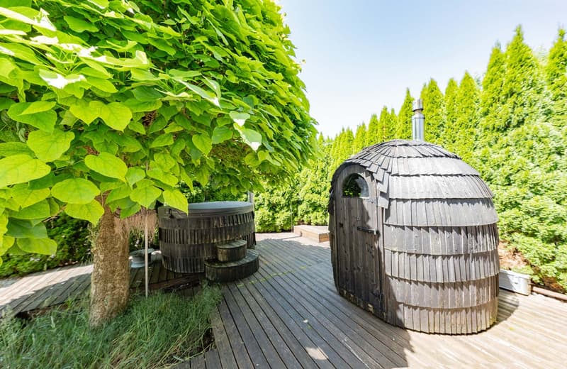 Wooden deck with a dome-shaped wooden structure, likely a sauna or outdoor shelter, surrounded by lush greenery including trees and shrubs.