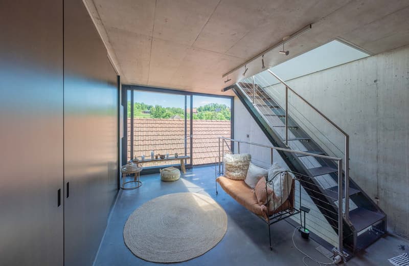 Spacious living room with concrete ceiling, large windows overlooking a balcony, modern furniture including a couch and coffee table, and a spiral staircase leading to an upper level.