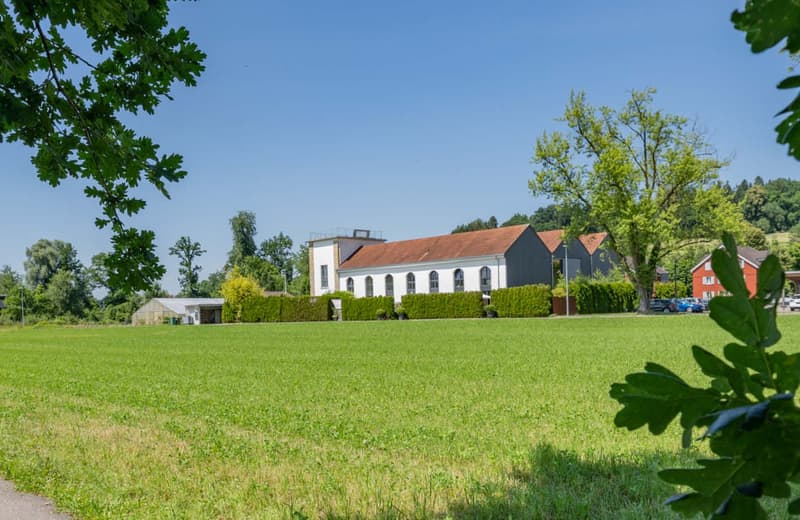 2-story building with a red tile roof, surrounded by a well-maintained lawn and lush greenery. The building has large windows and appears to be a residential or commercial property.