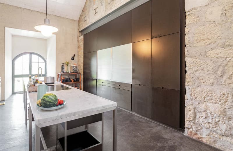 Modern kitchen with dark cabinets, marble countertop, kitchen island, and appliances including a dishwasher. The kitchen has exposed stone walls, giving it a rustic industrial feel.