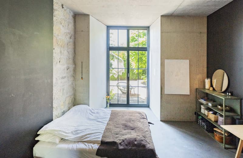 Spacious bedroom with large windows overlooking a lush, green outdoor view. The room features concrete walls, a bed with a patterned blanket, and a small shelf unit with decor items.