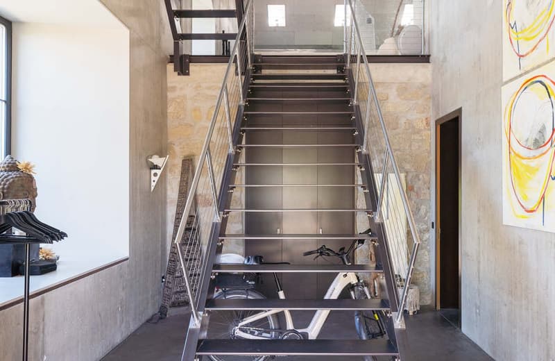 Metal staircase with open treads, leading to an upper level. The stairs have a modern, industrial design with a metal frame and glass railings. The space appears to be a loft or converted industrial space, with exposed concrete walls and artwork on the wa