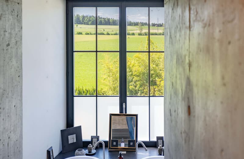 Large windows overlooking a scenic rural landscape with green fields and trees, concrete walls, framed artwork and decorative items on a table in the foreground