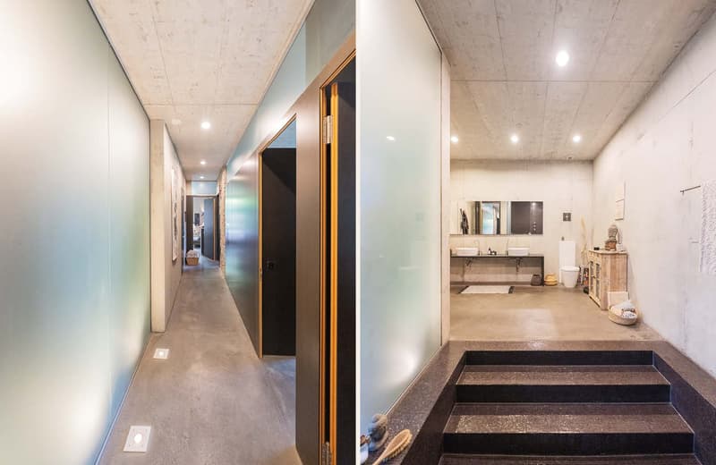 The image shows a long hallway with concrete walls and ceiling, and recessed lighting. The hallway leads to a bathroom area with a raised platform and a vanity with sinks. The bathroom has a modern, minimalist design.
