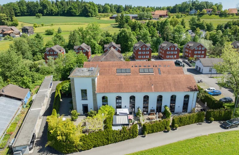 A large, multi-story building with a red tile roof surrounded by lush greenery and trees. The building appears to be a commercial or public facility, with several arched windows and a central entrance. There are several smaller residential buildings visib