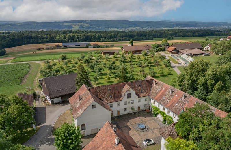 Large white house with red roof, surrounded by farmland, trees, and a fountain in the courtyard, aerial view