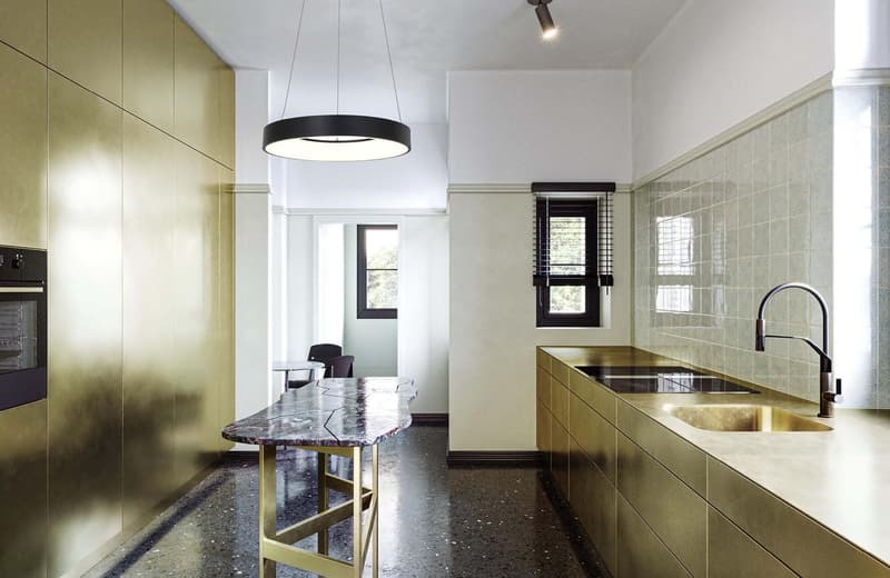 modern kitchen with brass-colored cabinets, white tiles, marble countertop, brass sink and faucet, large open windows, black ceiling light, modern black oven