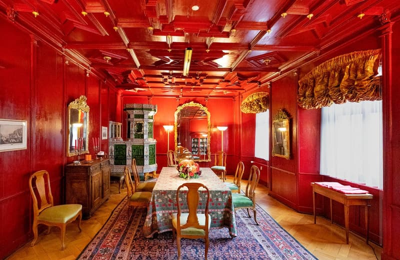Dining room with red walls, wooden floor, dining table, chairs, mirror, cabinet, flower arrangement, lamps, window with curtains