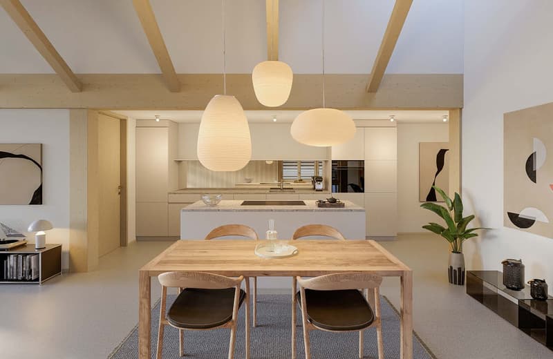 The image shows an open concept living space with a dining area and kitchen. The space features a wooden dining table with chairs, pendant lighting fixtures, and a modern kitchen with a central island. The room has a neutral color palette with wooden acce