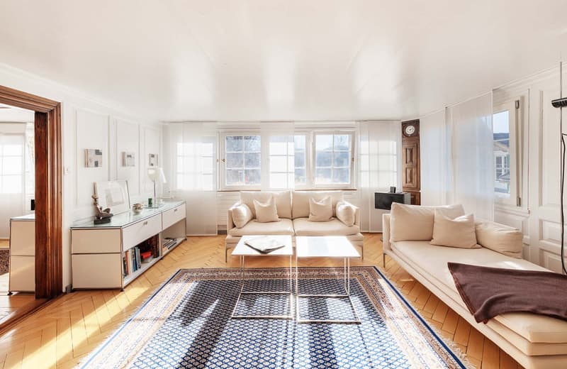 living room with two couches, two tables, rug, wooden floor, white walls, large windows, wooden door, cabinet, books, lamp, clock