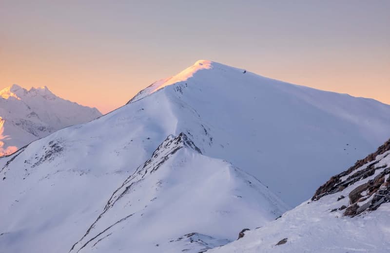 Snow-covered mountain peaks illuminated by a warm sunset glow, with rocky outcrops.