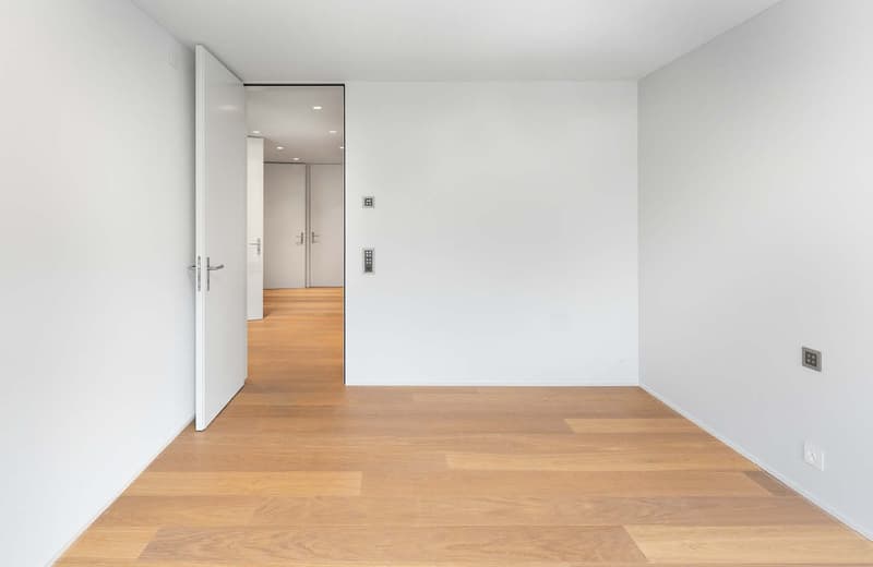 An empty room with hardwood floors, white walls, and an open door. There are two power outlets and a switch on the right wall.