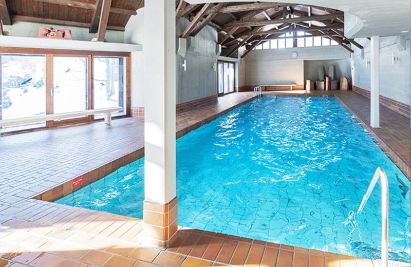 Two-lane indoor swimming pool, wooden ceiling beams, pool ladder, wooden benches, and glass windows.