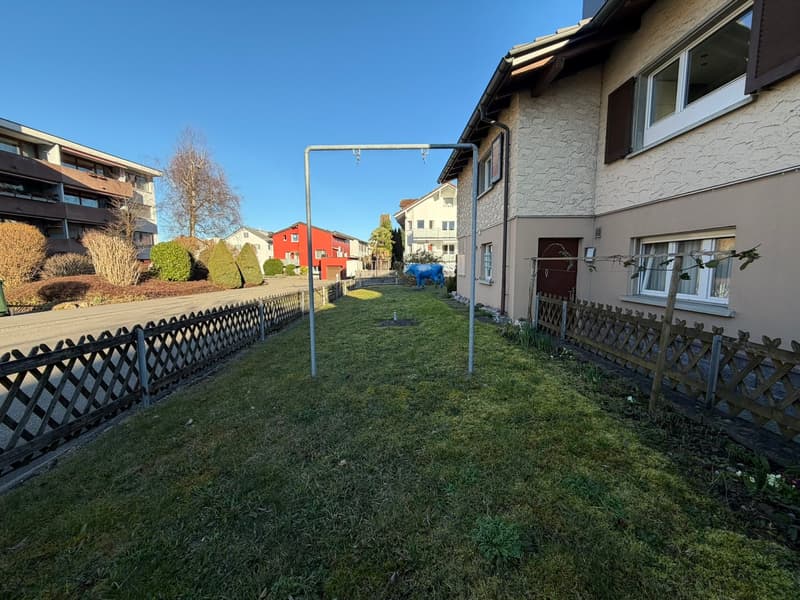 A small garden with a swing set, surrounded by a wooden fence, with a grassy lawn and a blue cow statue.