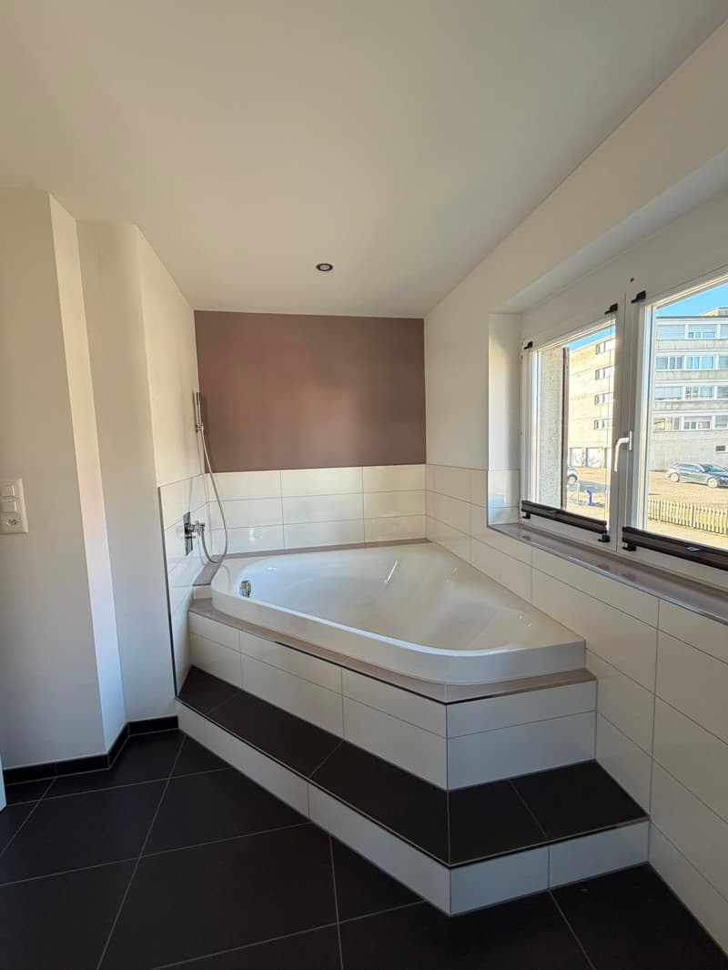 Bathroom with a large bathtub, white tiles, black floor, two windows, shower head