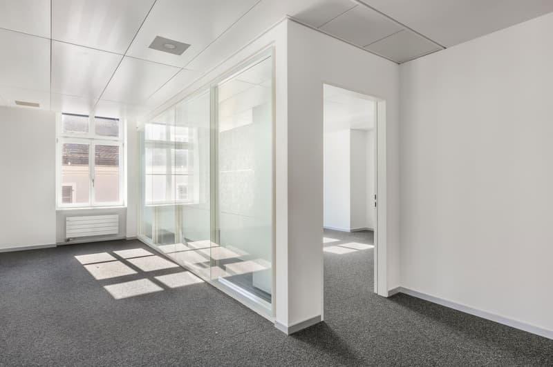 Large open office space with white walls, gray carpeted floor, and glass partition walls. The room has large windows providing natural light, and a doorway leading to another room.