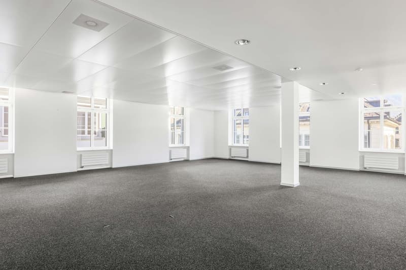 Large open office space with white walls, gray carpeted floor, and multiple windows providing natural light. The room has a clean, modern design with recessed lighting fixtures in the ceiling.
