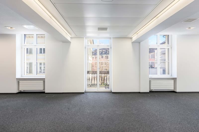 Large open office space with white walls, gray carpeted floor, and large windows on both sides providing natural light. The room has a high ceiling with recessed lighting. There are built-in cabinets or storage units along the walls.