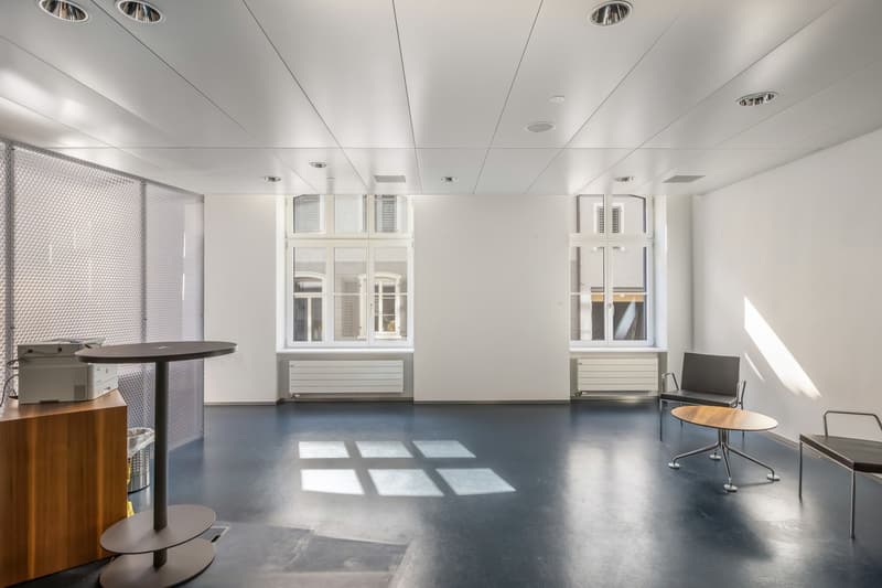 The image shows a modern, minimalist office space with white walls, a concrete floor, and large windows that let in natural light. The room has a round table, a couple of chairs, and some computer equipment. The space appears to be designed for a small of