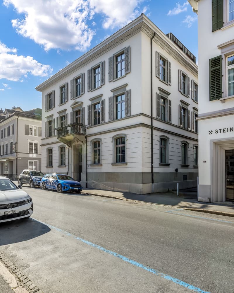 A multi-story building with a classical architectural style, featuring large windows, shutters, and a balcony. The building has a light-colored facade and is situated on a street with parked cars.