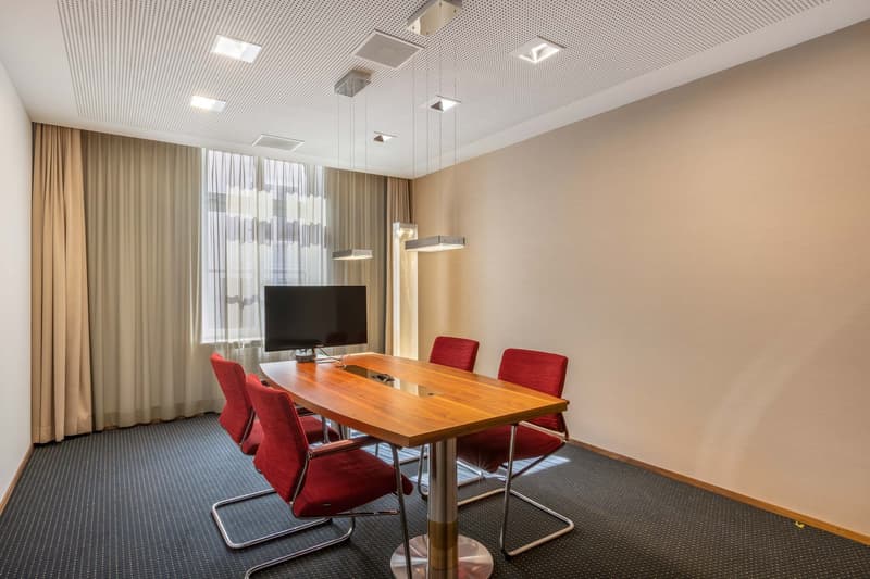 Spacious conference room with wooden table, red chairs, TV, and pendant lighting fixtures on the ceiling