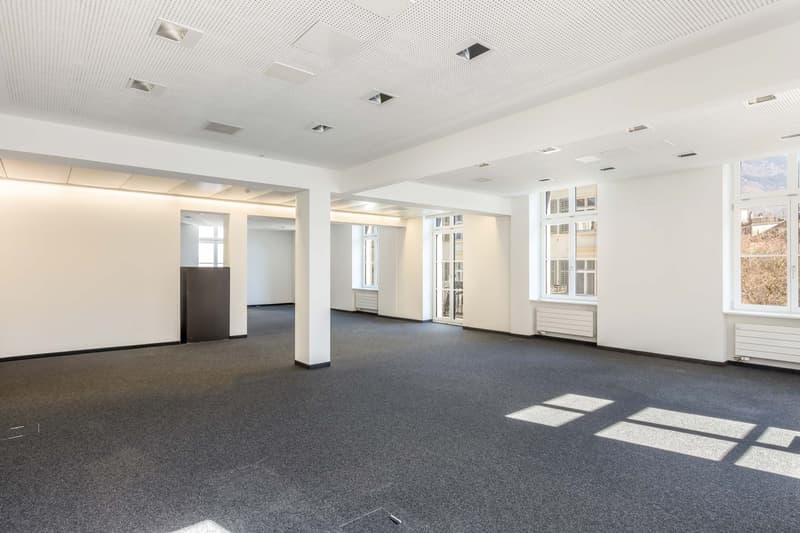 Large open office space with white walls, gray carpeted floor, and multiple windows providing natural light. The space appears to be empty and ready for use.