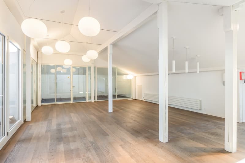 Large open-plan office space with hardwood floors, white walls, and multiple pendant light fixtures. The space features glass walls and doors, providing a bright and airy atmosphere. There appears to be a balcony or terrace visible through the glass walls