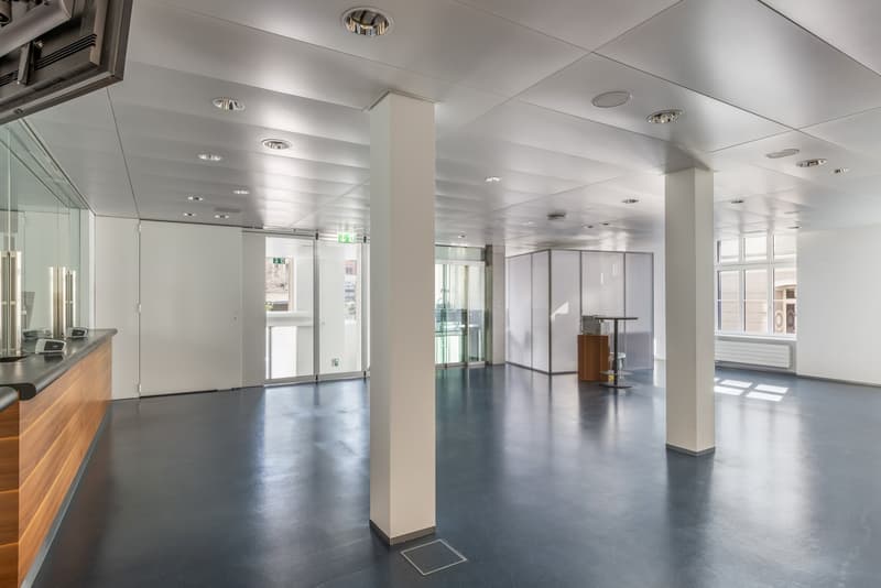 The image shows a large, open office space with a modern, minimalist design. The space features a reception area with a wooden counter, and the rest of the office is characterized by white walls, a gray concrete floor, and recessed lighting fixtures in th