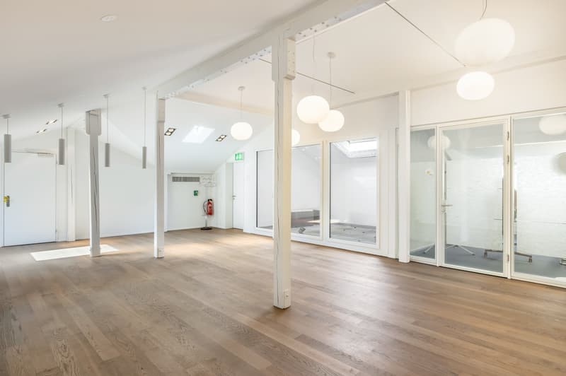 Large open-plan office space with hardwood floors, white walls, and large windows. The space features several pendant lights and columns, as well as a glass partition wall. The room appears to be well-lit and designed for a professional office or commerci