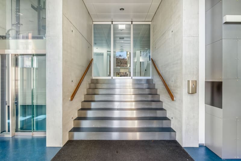 Spacious staircase with metal steps, glass walls, and wooden handrails leading to an upper level