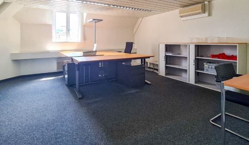 An empty office with two desks, two cabinets, chairs, and a radiator. A large window on the left side of the room. White ceiling and walls.