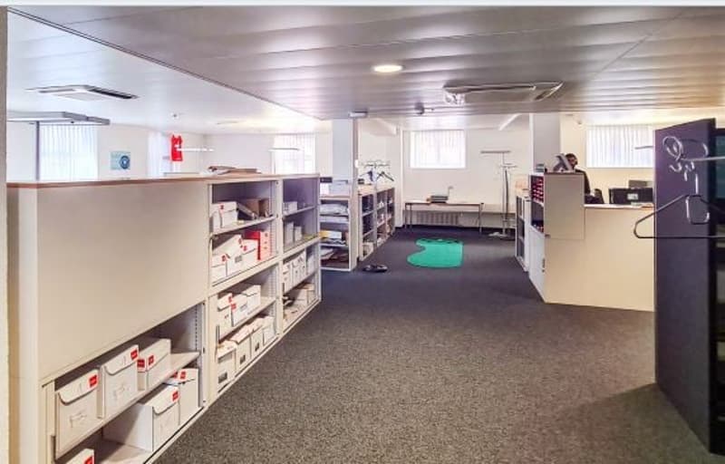 Office space with grey carpet, white walls, multiple shelves, desks, large windows, and fire extinguisher