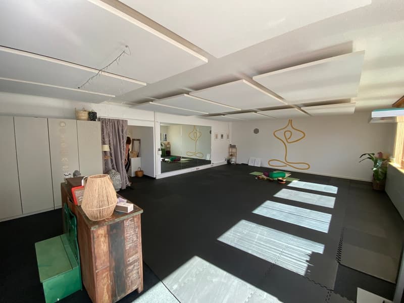 large open room with white walls, black matte floor, yoga mats, yoga objects, yoga mat, windows, plants, wooden desk, lamp